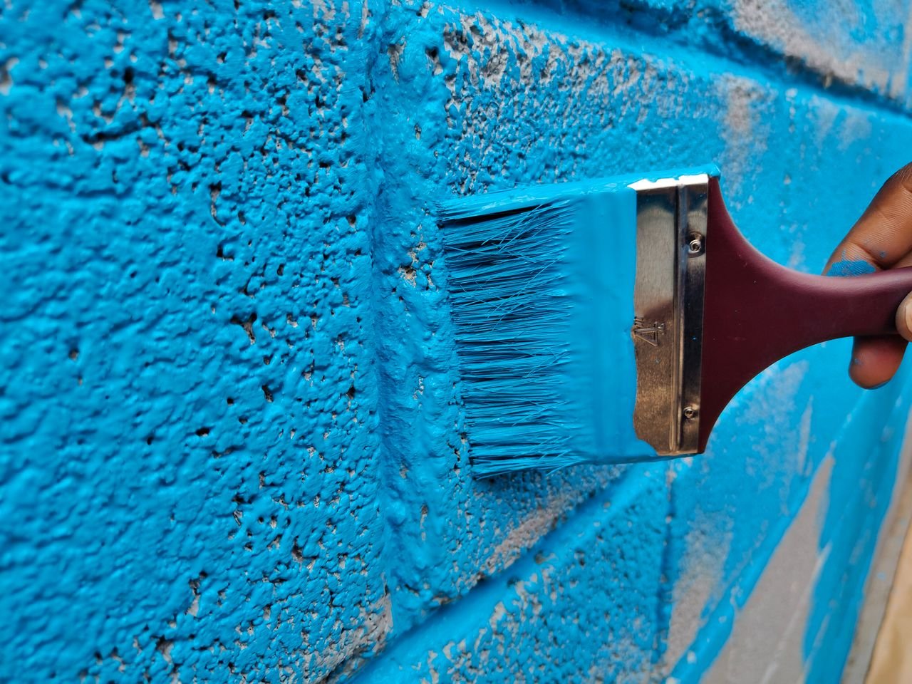A hand painting a brick wall with vibrant blue paint using a brush.
